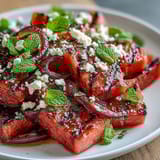 A colorful platter of grilled watermelon cubes topped with feta crumbles, red onion, and mint leaves, drizzled with balsamic glaze.