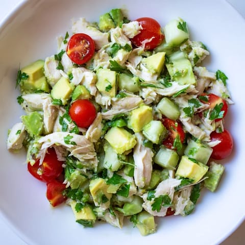 Freshly prepared avocado chicken salad served in a white bowl with cherry tomatoes and cucumber, perfect for a light lunch.  